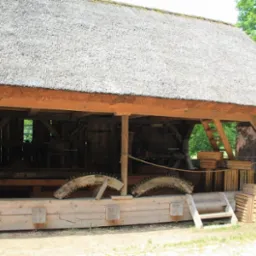 Drewniana konstrukcja z strzechą, widoczne elementy starego warsztatu, w tym fragmenty drewnianych kół zębatych.