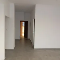 Wnętrze pustego, odnowionego pokoju z szarymi płytkami na podłodze i białymi ścianami. Widoczne otwarte drzwi do kolejnego pomieszczenia.