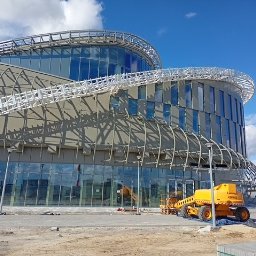 Kamil Milewski usługi Remontowo-Budowlane - Nowoczesny budynek biurowy z przeszkloną fasadą i metalową konstrukcją, w trakcie prac wykończeniowych. Na pierwszym planie żółty podnośnik i materiały budowlane.