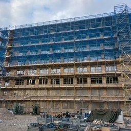 Kamil Milewski usługi Remontowo-Budowlane - Wielopiętrowy budynek w trakcie prac elewacyjnych, otoczony rusztowaniami z niebieską siatką ochronną. Widoczne okna i materiały budowlane na placu.