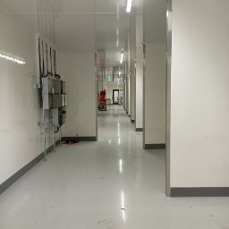 Kamil Milewski usługi Remontowo-Budowlane - Długi, jasny korytarz z białymi ścianami i podłogą, oświetlony lampami sufitowymi. Widoczne instalacje elektryczne i wózek widłowy w oddali. Minimalistyczny, sterylny wygląd.