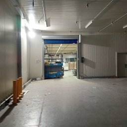 Kamil Milewski usługi Remontowo-Budowlane - Wnętrze hali produkcyjnej z widocznym podnośnikiem nożycowym, metalowymi panelami ściennymi i oświetleniem sufitowym. Widok na otwartą przestrzeń magazynową.