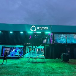 Kamil Milewski usługi Remontowo-Budowlane - Dwukondygnacyjny pawilon z kontenerów IQOS na zielonej trawie, oświetlony neonowym światłem. Mężczyzna stoi obok. Schody prowadzą na górę. Na dachu logo IQOS.