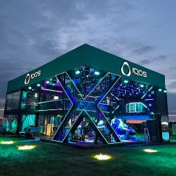 Kamil Milewski usługi Remontowo-Budowlane - Dwukondygnacyjny pawilon z logo IQOS, oświetlony neonami w kształcie litery X, na trawiastym terenie o zmierzchu. Widoczne osoby i pojazdy w tle.