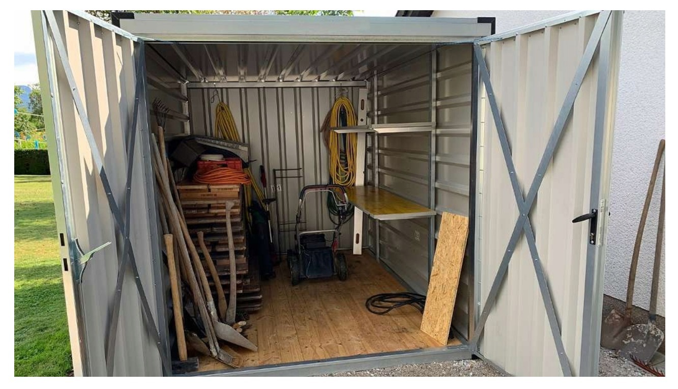 Otwarty metalowy magazyn ogrodowy z narzędziami, wewnątrz widoczna kosiarka, deski i półki, na zewnątrz łopaty opierają się o ścianę.