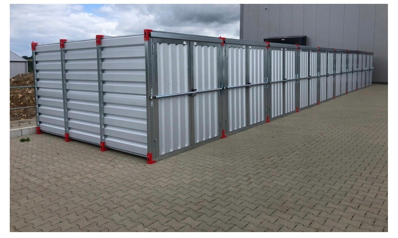 Zestaw modułowych, metalowych boksów magazynowych z czerwonymi narożnikami, ustawionych na brukowanym placu przy szarym budynku.