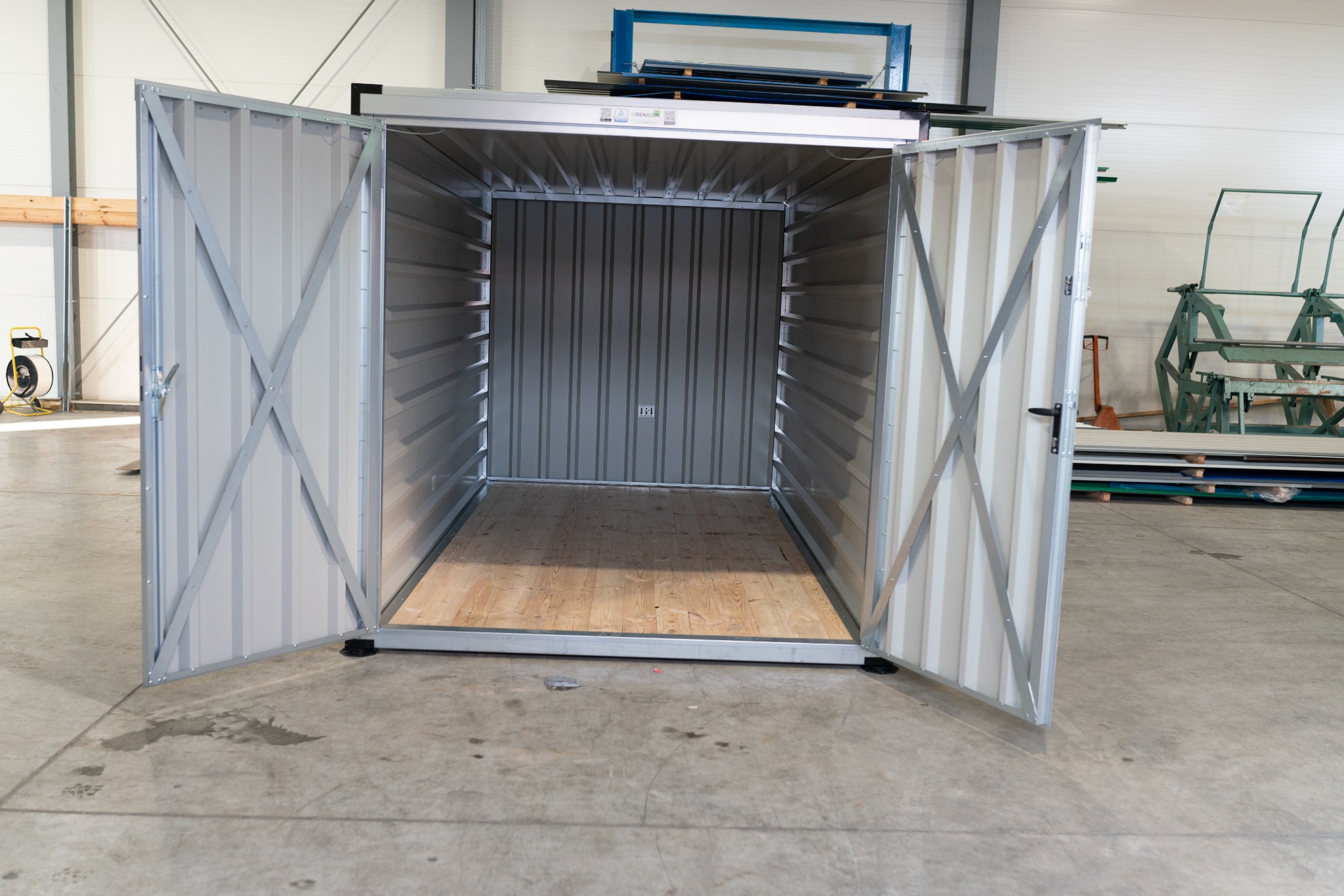 Otwarty, metalowy kontener magazynowy z drewnianą podłogą, widok z przodu, w tle hala produkcyjna.