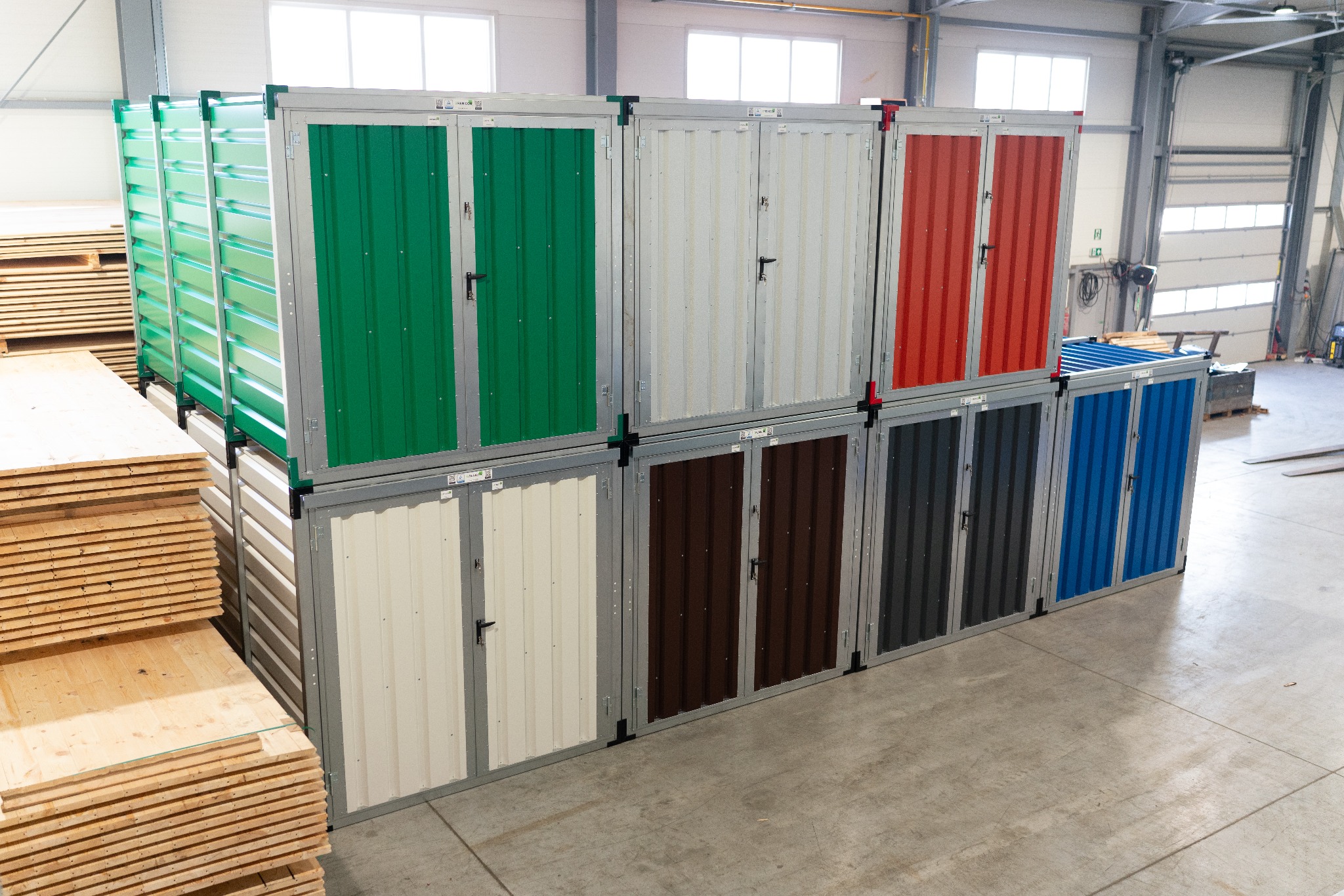 Rząd kolorowych, metalowych kontenerów magazynowych zamykanych na klucz, ustawionych w hali produkcyjnej, obok sterty drewnianych desek.