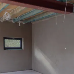 Surowe wnętrze z oknem, ściany pokryte świeżym, szarym tynkiem, widoczne elementy konstrukcyjne sufitu i zwisające przewody elektryczne.