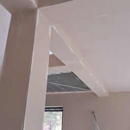 VitalTynki - Gładkie, jasne ściany i sufit po pracach wykończeniowych. Widoczne filary i fragment okna z widokiem na zieleń. Kabel wystaje ze ściany.