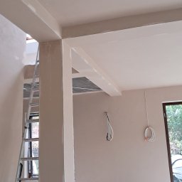 VitalTynki - Wnętrze w trakcie prac wykończeniowych: świeżo położone tynki na ścianach i suficie, widoczna drabina, kable i wiadro na podłodze. Jasne, naturalne światło wpada przez okno.