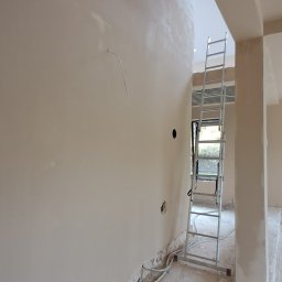 VitalTynki - Ściana po nałożeniu gładzi, widoczna drabina i przewody przy podłodze. Świeżo wykończone powierzchnie w jasnym odcieniu beżu, przygotowane do dalszych prac wykończeniowych.