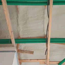 Poddasze z drewnianą konstrukcją, pokryte białą pianką izolacyjną, z widocznymi zielonymi rurami wentylacyjnymi i fragmentem sufitu podwieszanego.