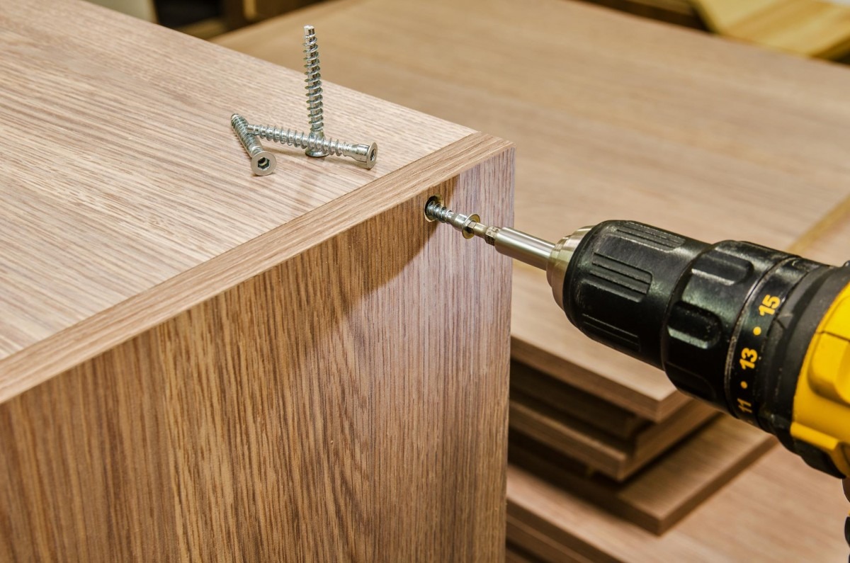 Skręcanie drewnianego elementu mebla wkrętarką, widoczne wkręty na blacie, obok stos płyt meblowych.
