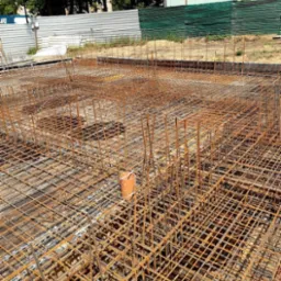 Siatka zbrojeniowa fundamentu z wystającymi prętami pionowymi i pomarańczową rurą wewnątrz, widok z góry na placu budowy z ogrodzeniem w tle.