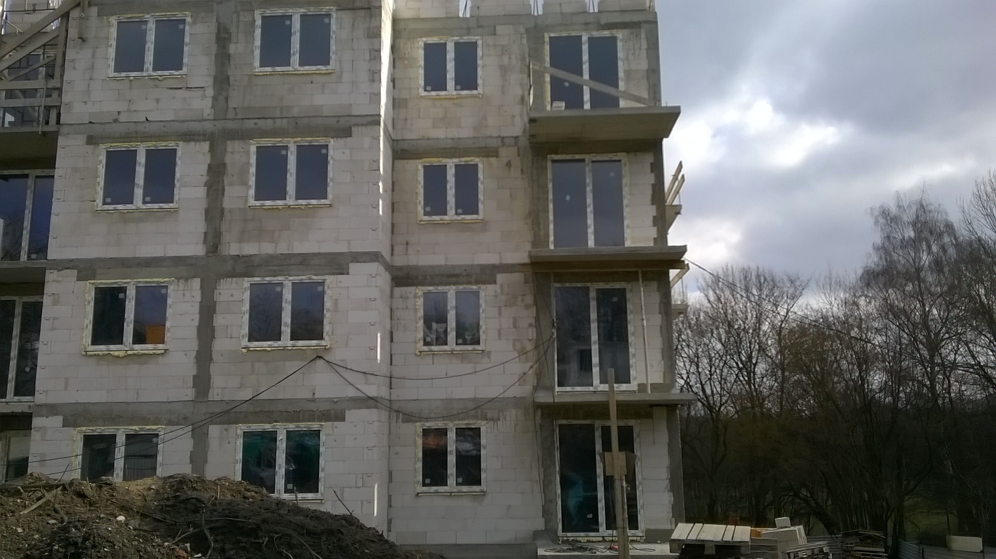 Wielopiętrowy budynek w trakcie budowy z bloczków, z zamontowanymi oknami i widocznym zbrojeniem oraz balkonami w surowym stanie, na tle pochmurnego nieba i drzew bez liści.
