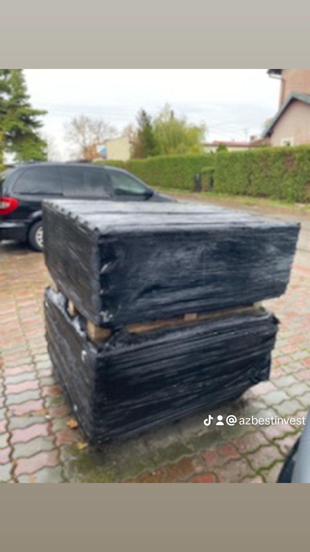 Dwie palety blachy w czarnym opakowaniu foliowym, ułożone jedna na drugiej, widoczne na zewnątrz, obok zaparkowany samochód.