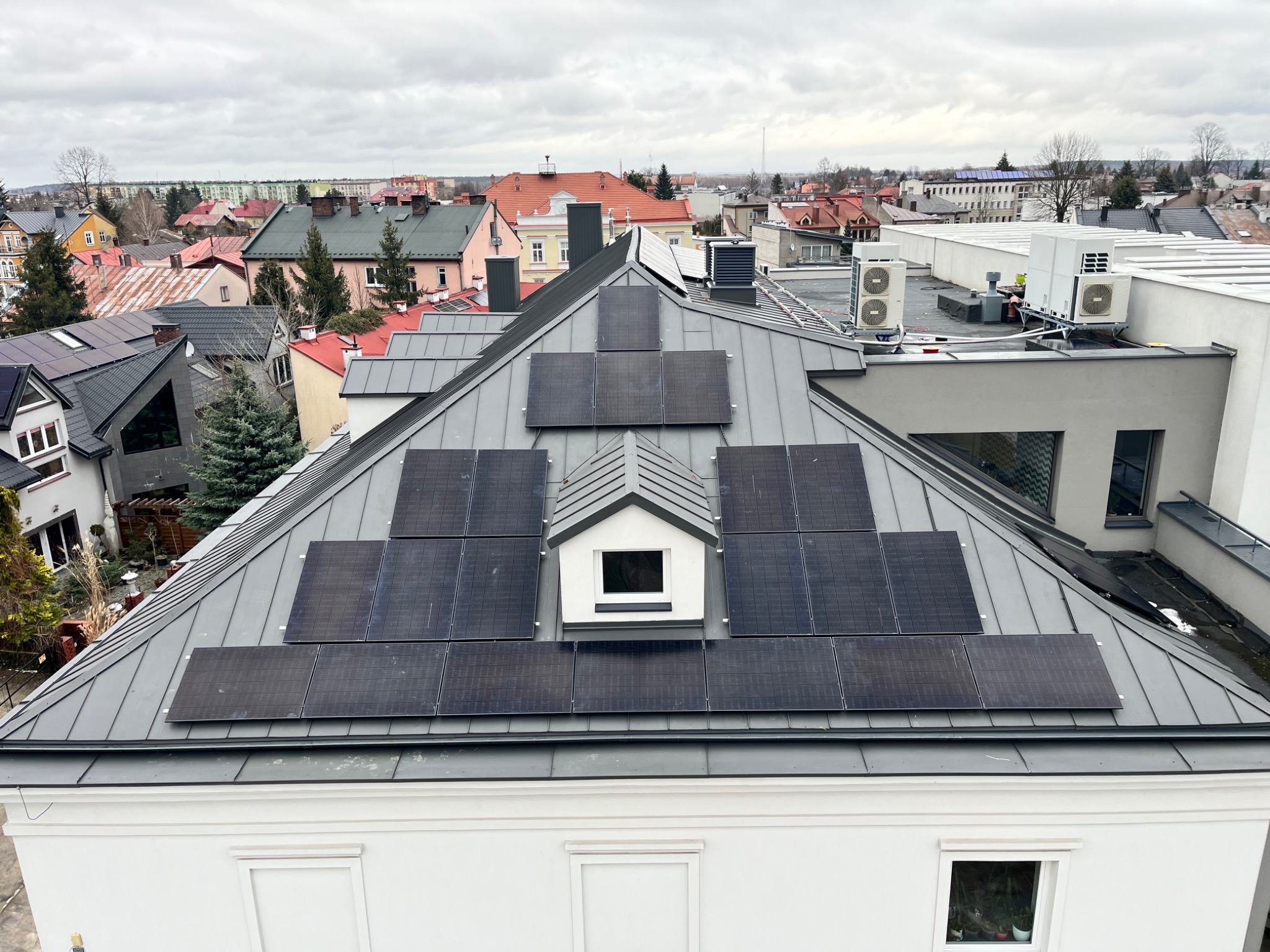 Widok z góry na szary dach z panelami słonecznymi ułożonymi symetrycznie wokół lukarny, w tle zabudowa miejska i pochmurne niebo.