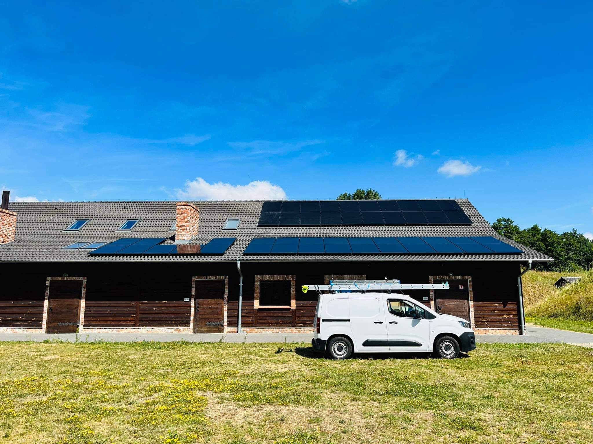 Instalacja paneli słonecznych na dachu budynku z ceglanym kominem, biały samochód dostawczy z drabiną oparty o ścianę budynku, słoneczny dzień.