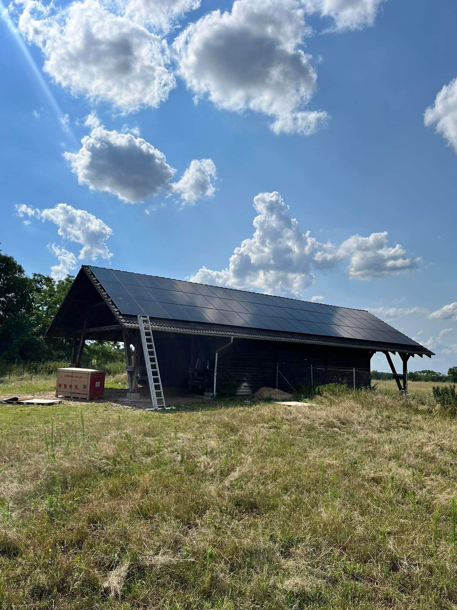 Ciemna, drewniana stodoła z zamontowanymi panelami fotowoltaicznymi na dachu, z boku oparta drabina, w tle błękitne niebo z białymi chmurami, na ziemi kartony z logo firmy fotowoltaicznej.