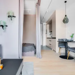 Jasne wnętrze małego mieszkania z wydzieloną strefą sypialnianą za pomocą zasłon, widoczny aneks kuchenny, stół z krzesłami i sofa z dekoracjami.