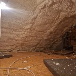 RBBUD Roman Bimkiewicz - Poddasze po ociepleniu pianką PUR, widoczne fragmenty piany na podłodze i ścianach. Kabel elektryczny i fragment lampy oświetlają przestrzeń.