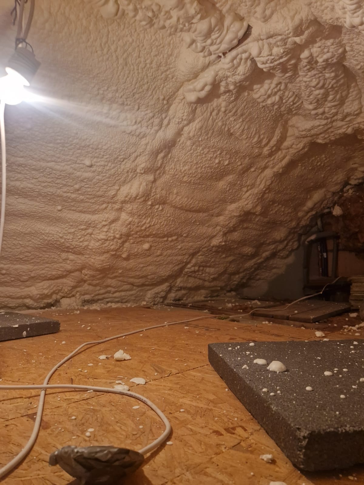 Poddasze po ociepleniu pianką PUR, widoczne fragmenty piany na podłodze i ścianach. Kabel elektryczny i fragment lampy oświetlają przestrzeń.