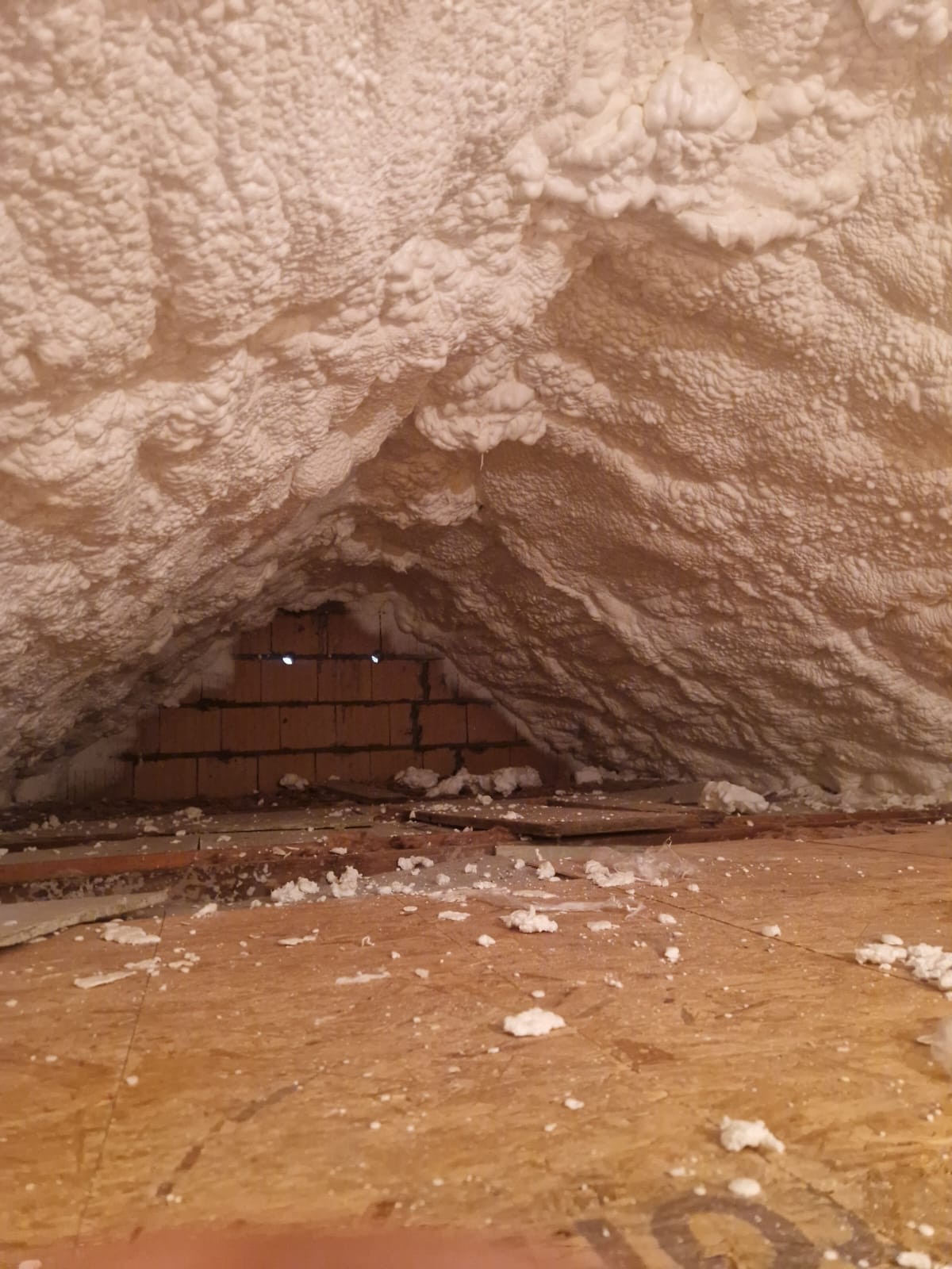 Poddasze po ociepleniu pianką PUR, widoczne cegły i drewniana podłoga z resztkami pianki. Tekstura pianki przypomina gęstą, nieregularną strukturę.