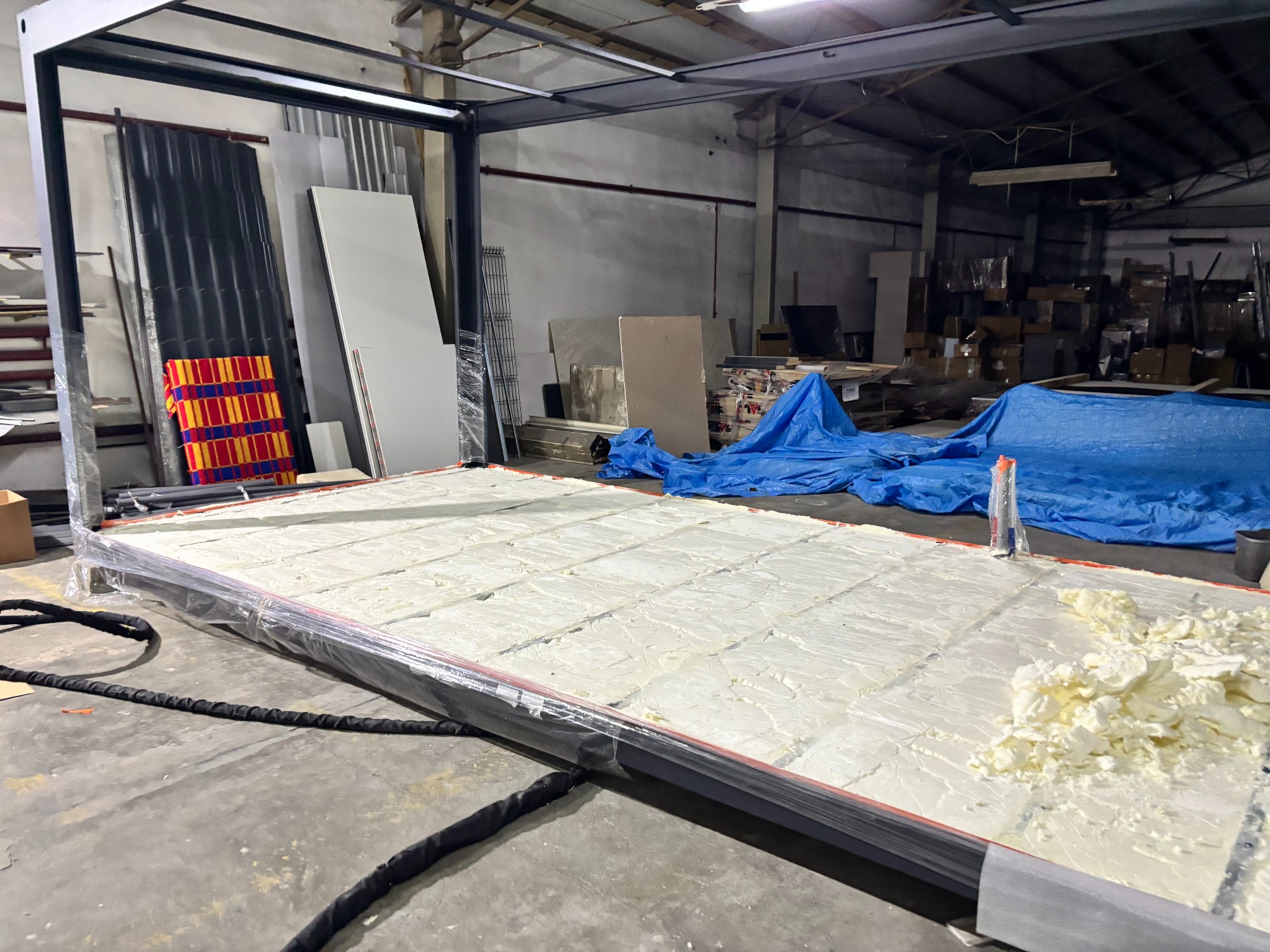 Proces aplikacji białej pianki izolacyjnej na metalową konstrukcję w hali. Widoczne materiały budowlane i narzędzia w tle.