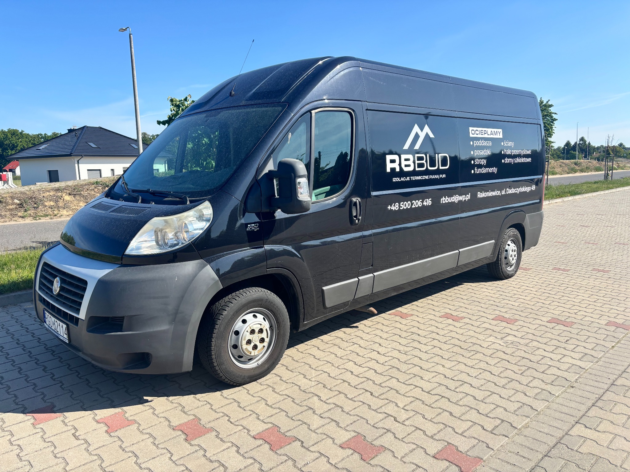Czarny bus firmy RBBUD z reklamą ocieplania pianką PUR. Na pojeździe widoczne logo, numer telefonu i adres e-mail. W tle budynek mieszkalny i błękitne niebo.