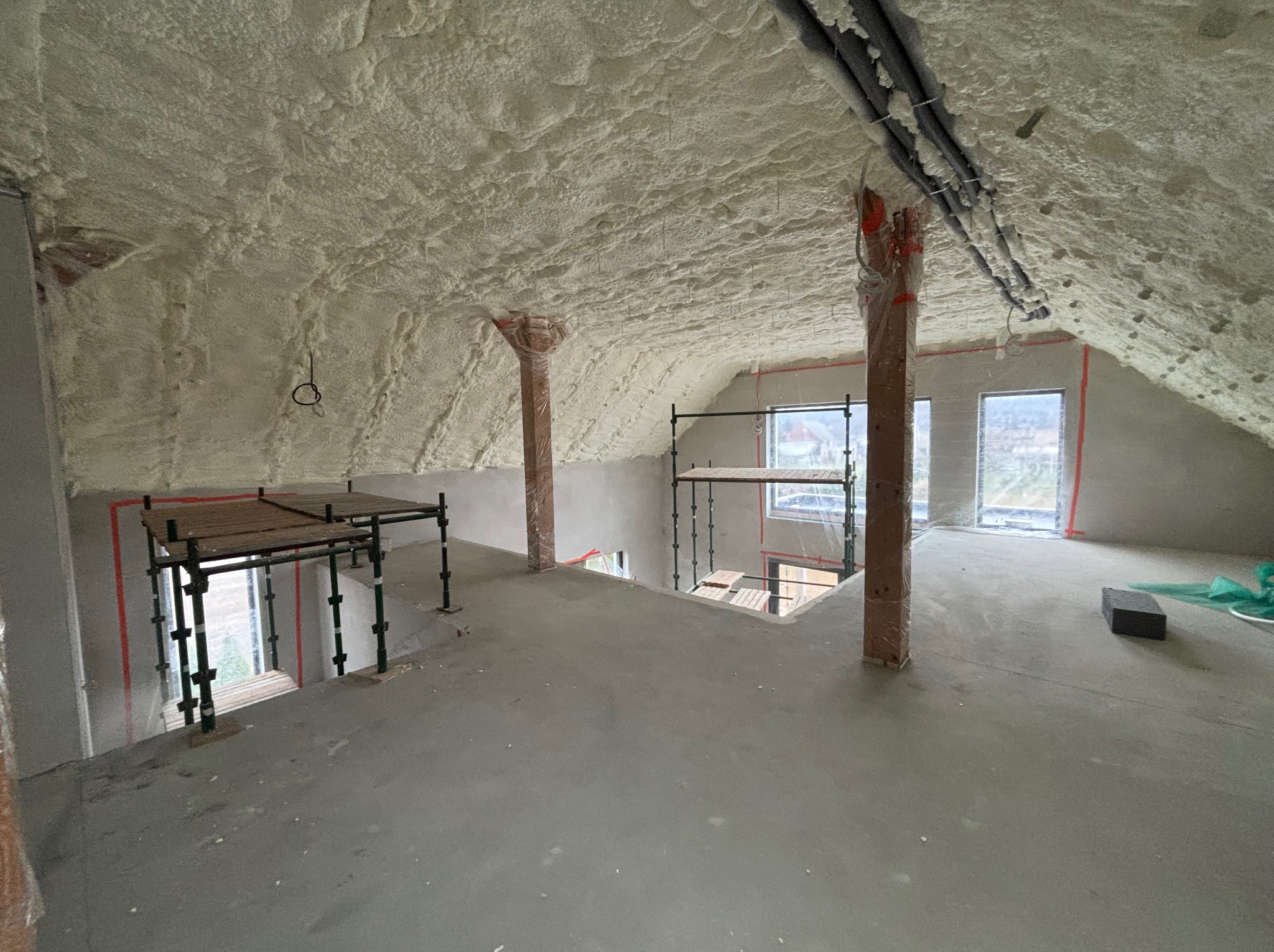 Poddasze po ociepleniu pianką PUR, widoczne rusztowania i zabezpieczone folią filary. Ściany i sufit pokryte grubą warstwą piany, rury pod sufitem również zaizolowane.