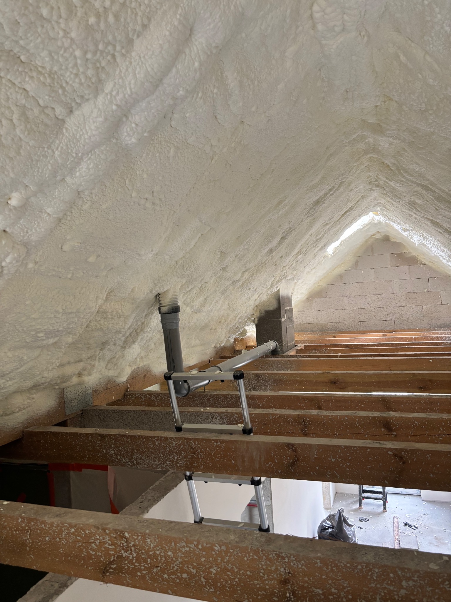 Poddasze z widoczną izolacją pianką PUR na skośnych ścianach i suficie, drewniane belki stropowe, fragment instalacji wentylacyjnej i drabina w tle.