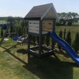 Zestaw zabaw dla dzieci na trawniku, z drewnianą wieżą z daszkiem, zjeżdżalnią i huśtawką, pomalowany na szaro i biało, w tle zielone drzewa i niebieskie niebo.