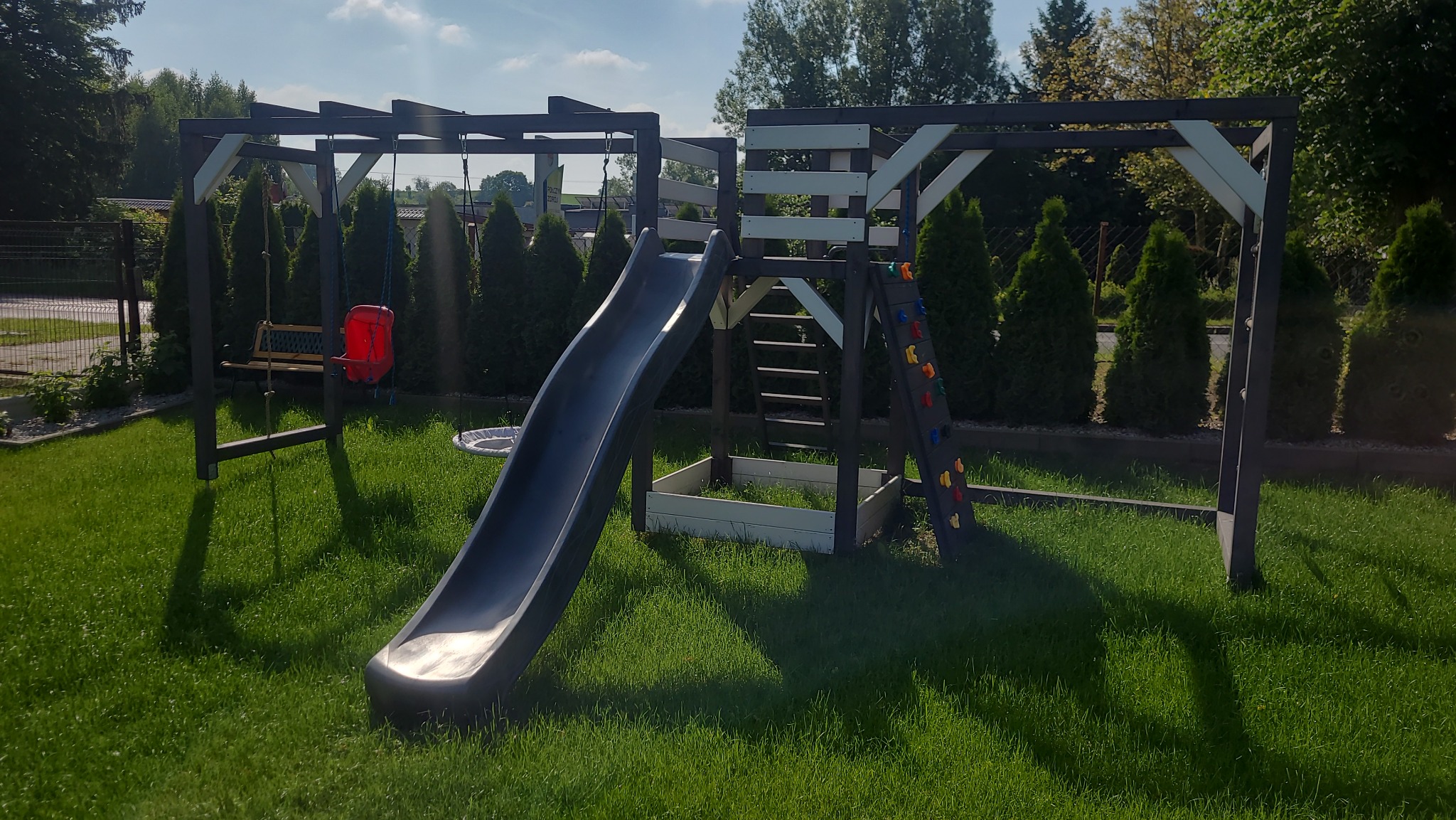 Kompleksowy plac zabaw z huśtawką, zjeżdżalnią, ścianką wspinaczkową i piaskownicą, wykonany z ciemnego drewna z białymi akcentami, na tle zielonego trawnika i żywopłotu.