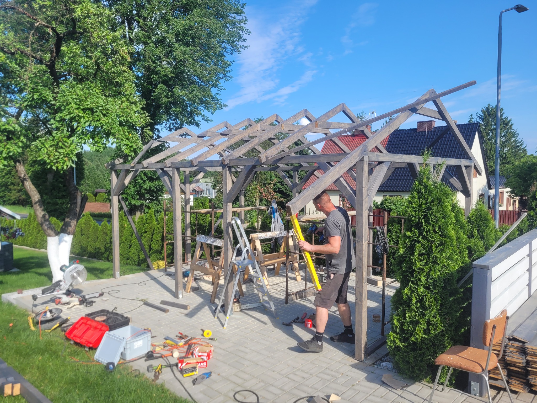 Montaż szkieletu drewnianej konstrukcji w ogrodzie, widoczne narzędzia, drabiny i pracownik z poziomicą na tle zieleni i domów.