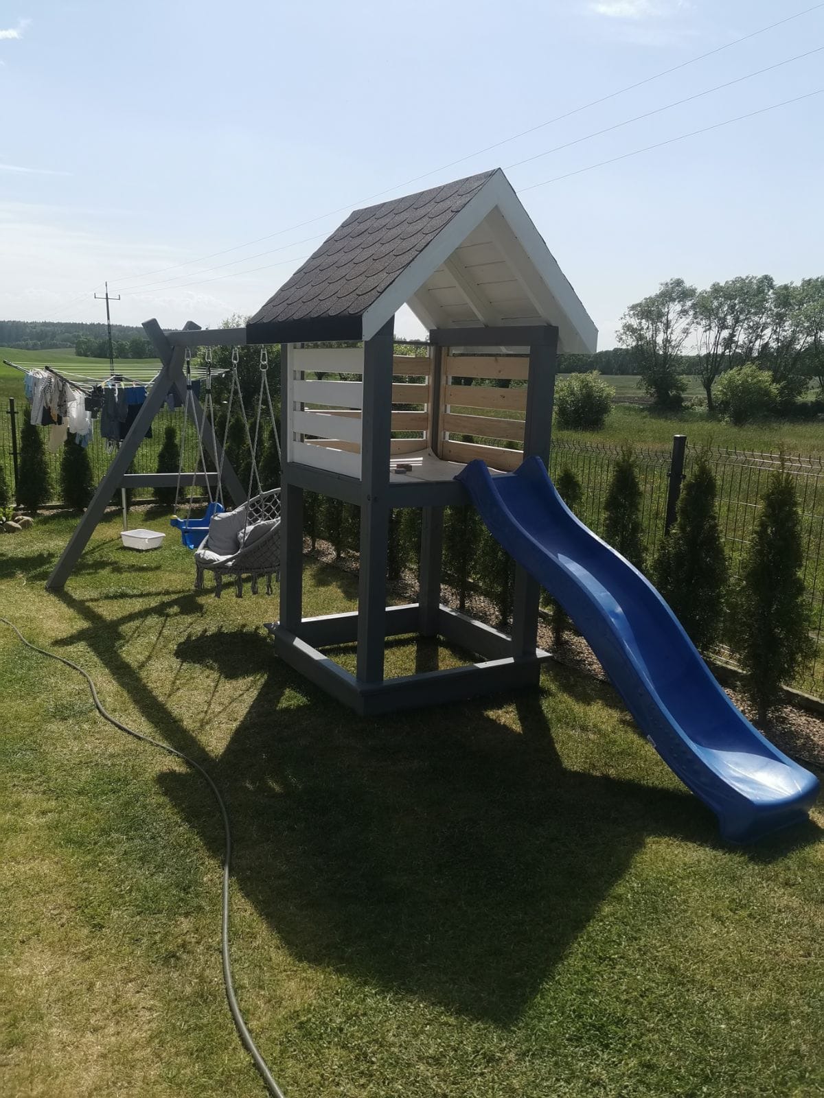 Drewniany plac zabaw z huśtawkami i zjeżdżalnią w Nowym Worowie, widoczny na tle zielonego trawnika i ogrodzenia z siatki.