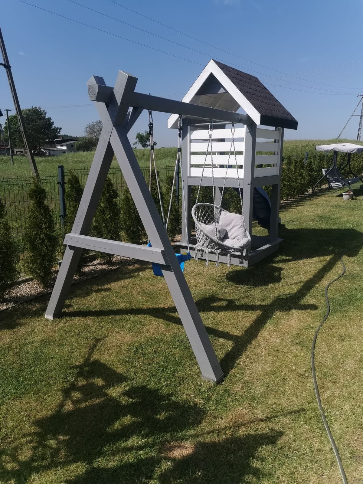 Szaro-biały plac zabaw z huśtawką typu bocianie gniazdo i zjeżdżalnią, umieszczony na trawniku w ogrodzie, widoczny przewód elektryczny na trawie.
