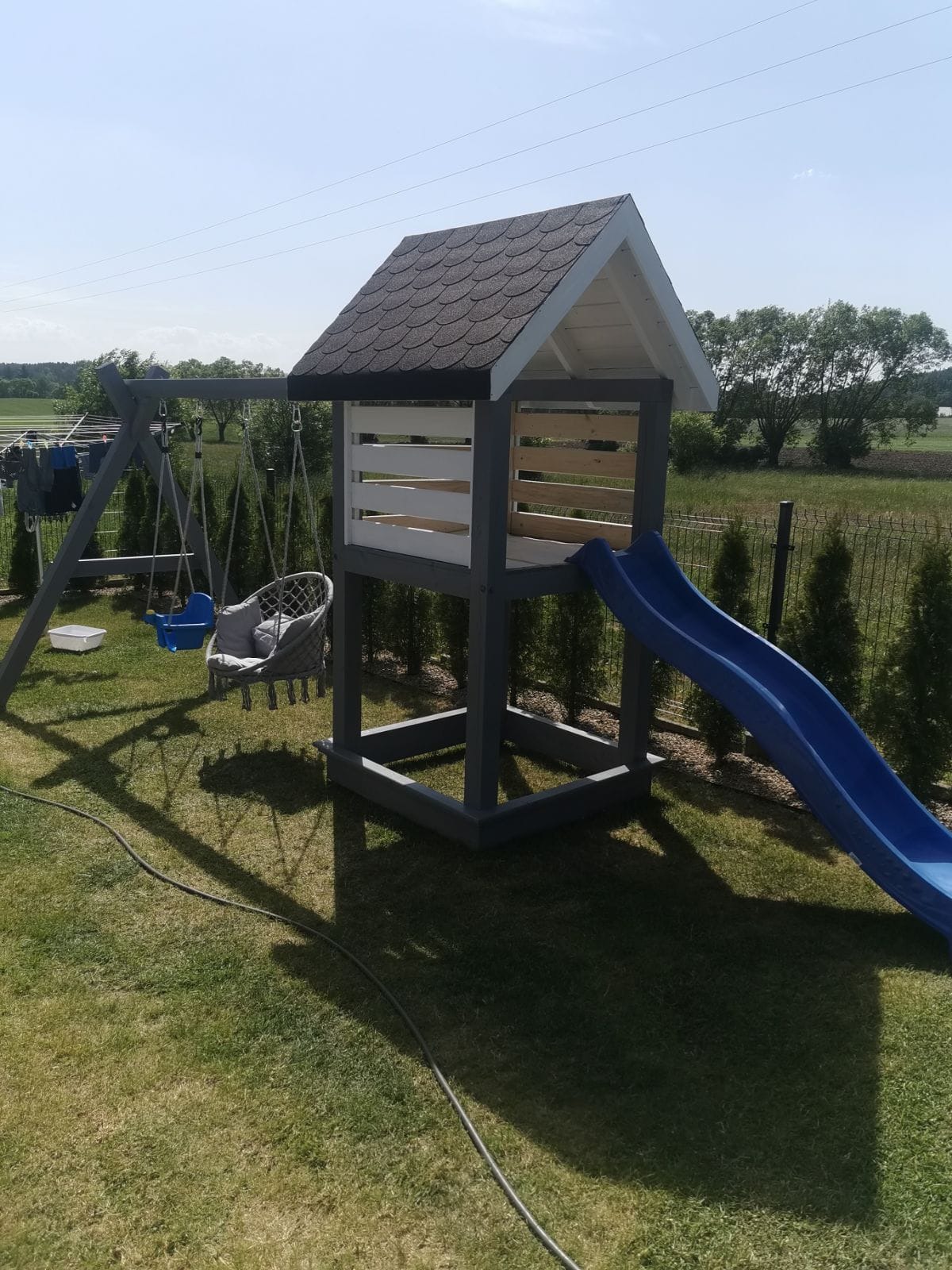 Zestaw zabaw dla dzieci na trawniku, z drewnianą wieżą z daszkiem, zjeżdżalnią i huśtawką, pomalowany na szaro i biało, w tle zielone drzewa i niebieskie niebo.