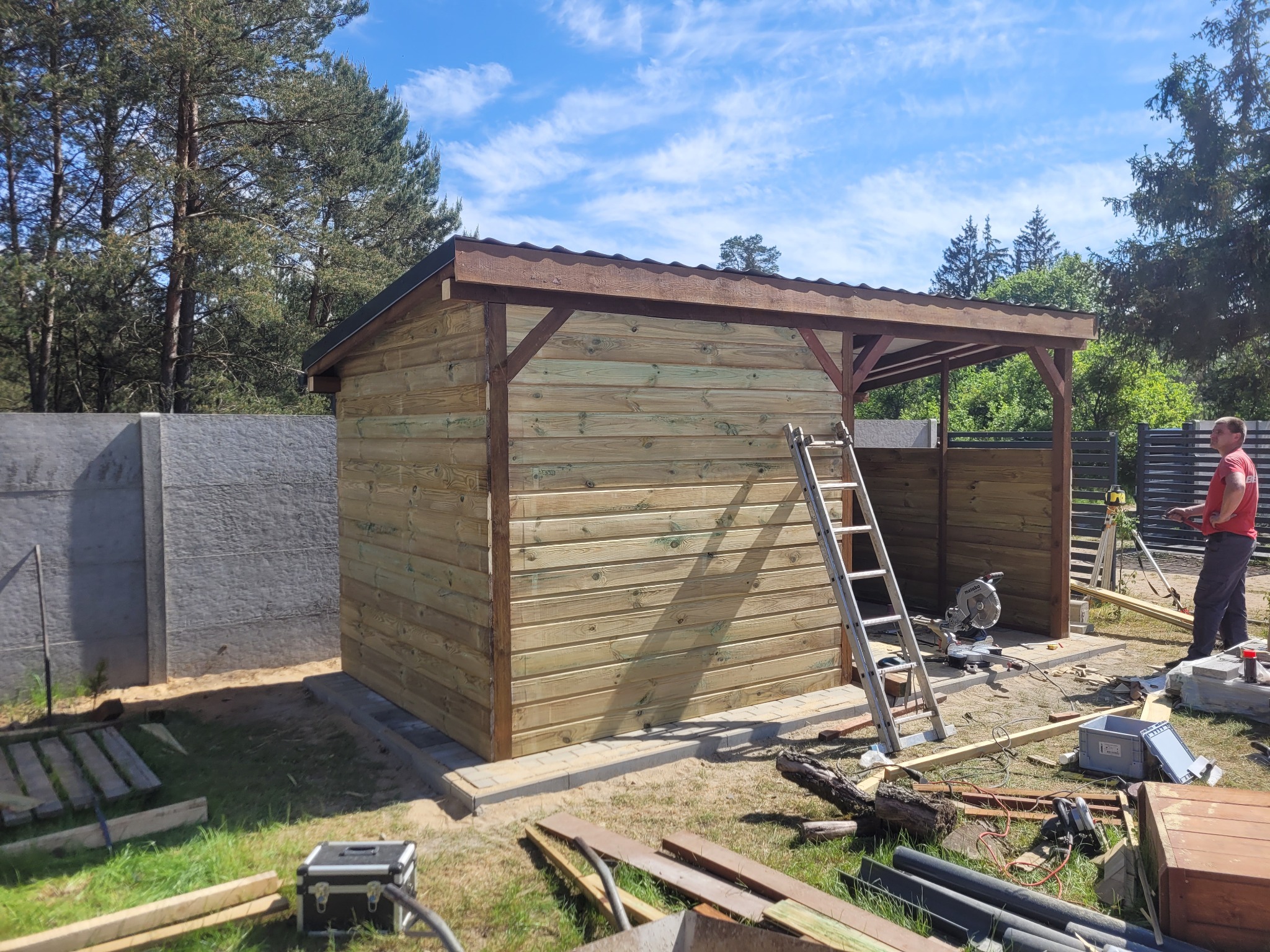 Budowa drewnianej altany z widoczną konstrukcją szkieletową, deskami elewacyjnymi w trakcie montażu i narzędziami pracy na trawie.