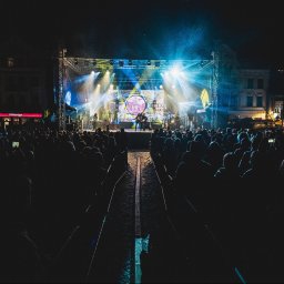 IMPRESION sp. z o.o. - Koncert plenerowy nocą: zespół na scenie oświetlonej reflektorami, tłum widzów w tle, budynki miejskie w oddali.