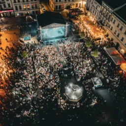 Nocny widok z lotu ptaka na koncert na rynku miejskim, tłum ludzi przed sceną, oświetlona fontanna, budynki w tle.