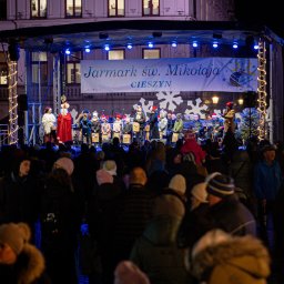 IMPRESION sp. z o.o. - Scena na Jarmarku św. Mikołaja w Cieszynie, na której występują dzieci w czerwonych czapkach i orkiestra dęta, oświetlona niebieskimi reflektorami, przed sceną zgromadzony tłum widzów.