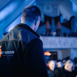 IMPRESION sp. z o.o. - Osoba w czarnej bluzie z logo firmy MP RION, stojąca tyłem do kamery, obserwuje scenę z widowni podczas wydarzenia.