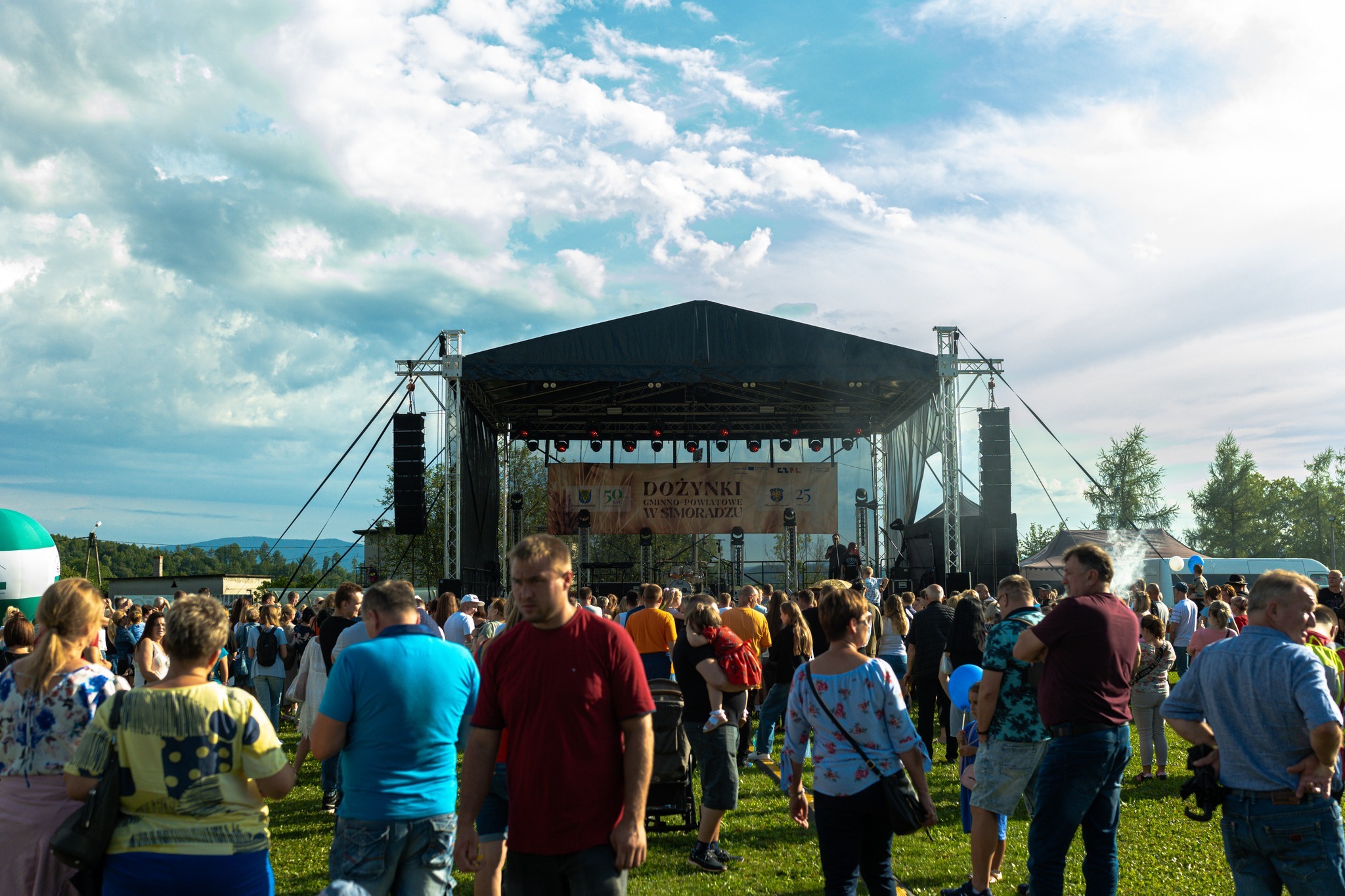 Scena plenerowa z nagłośnieniem i oświetleniem podczas dożynek gminnych, widoczny tłum ludzi i baner z napisem 'Dożynki Gminno Powiatowe w Simoradzu'.