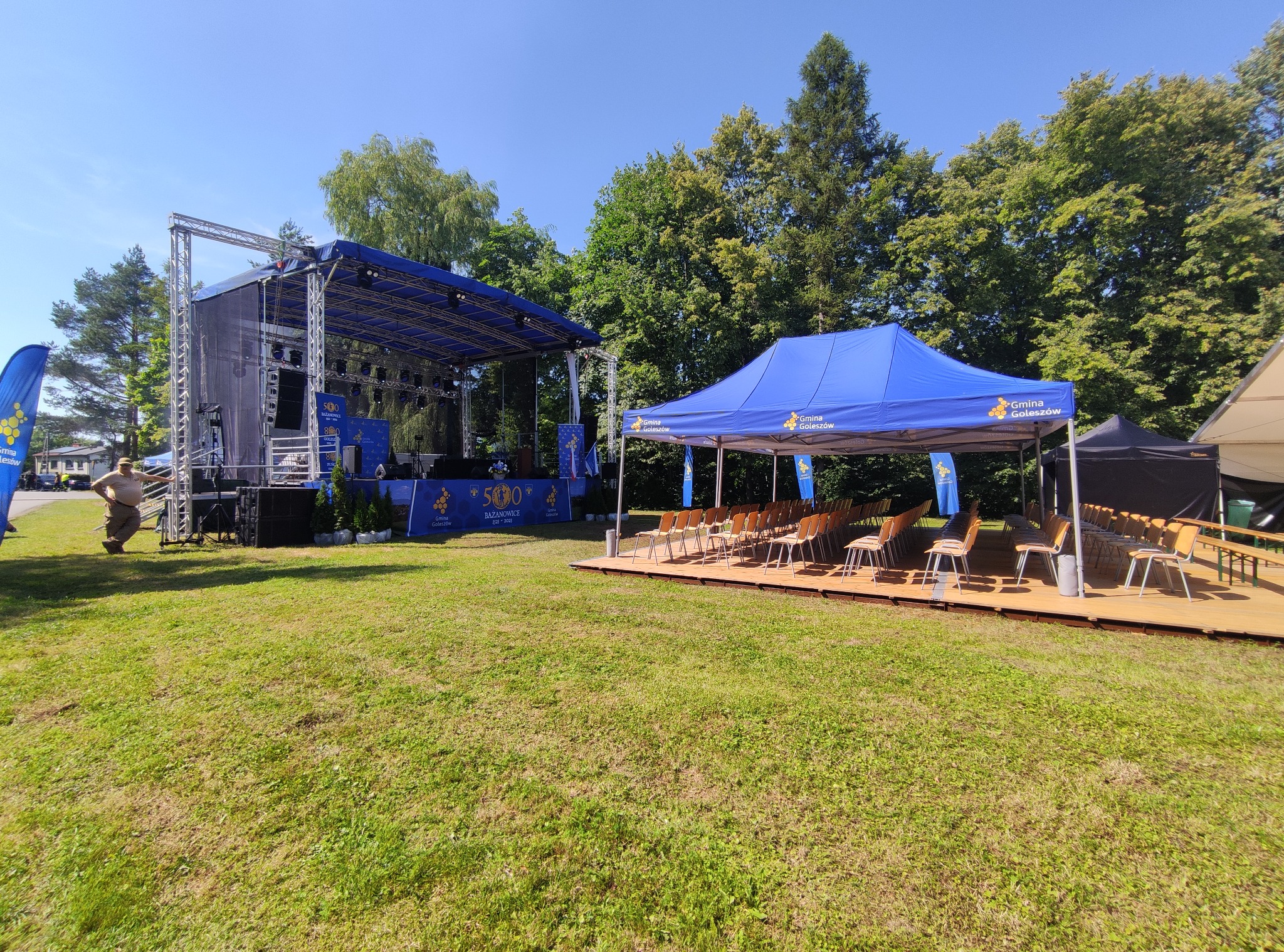 Scena plenerowa z konstrukcją stalową, nagłośnieniem i oświetleniem, obok namiot z krzesłami na drewnianym podeście, przygotowany na imprezę plenerową w słoneczny dzień.