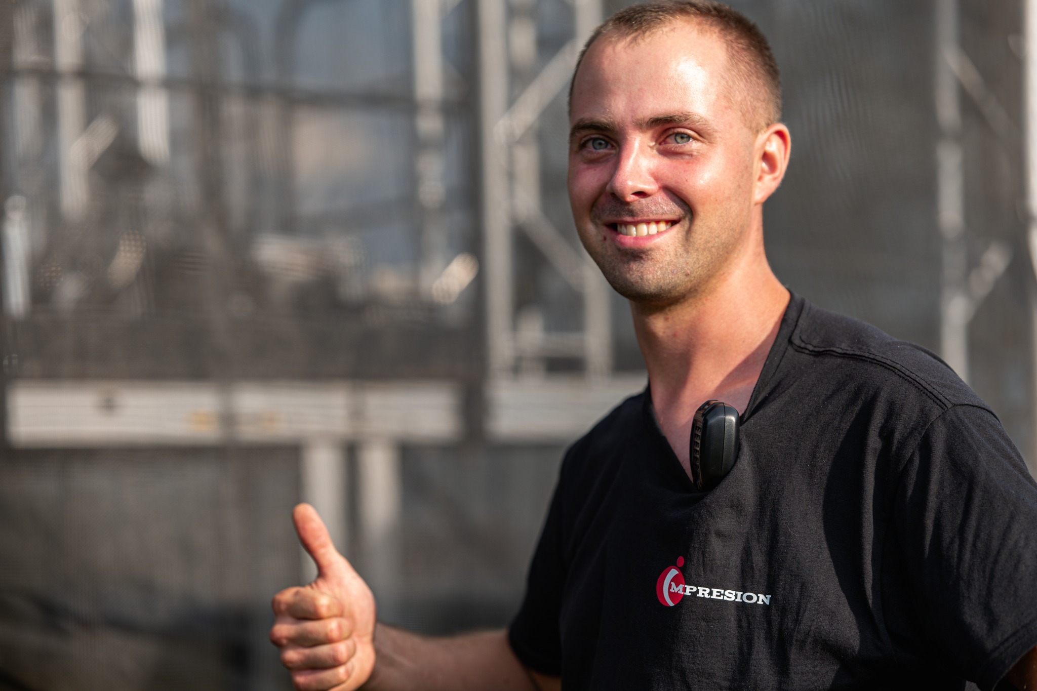 Uśmiechnięty mężczyzna w czarnej koszulce z logo firmy na tle konstrukcji scenicznej, trzymający kciuk w górę i z radiotelefonem przypiętym do koszulki.