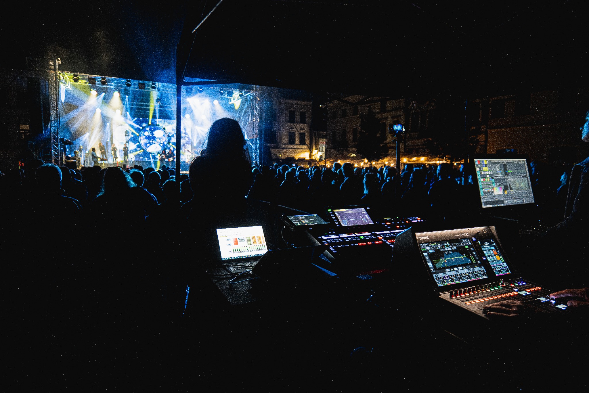 Koncert plenerowy nocą: widok zza konsolety oświetleniowej i dźwiękowej na scenę z występującymi muzykami i publicznością, oświetloną reflektorami.