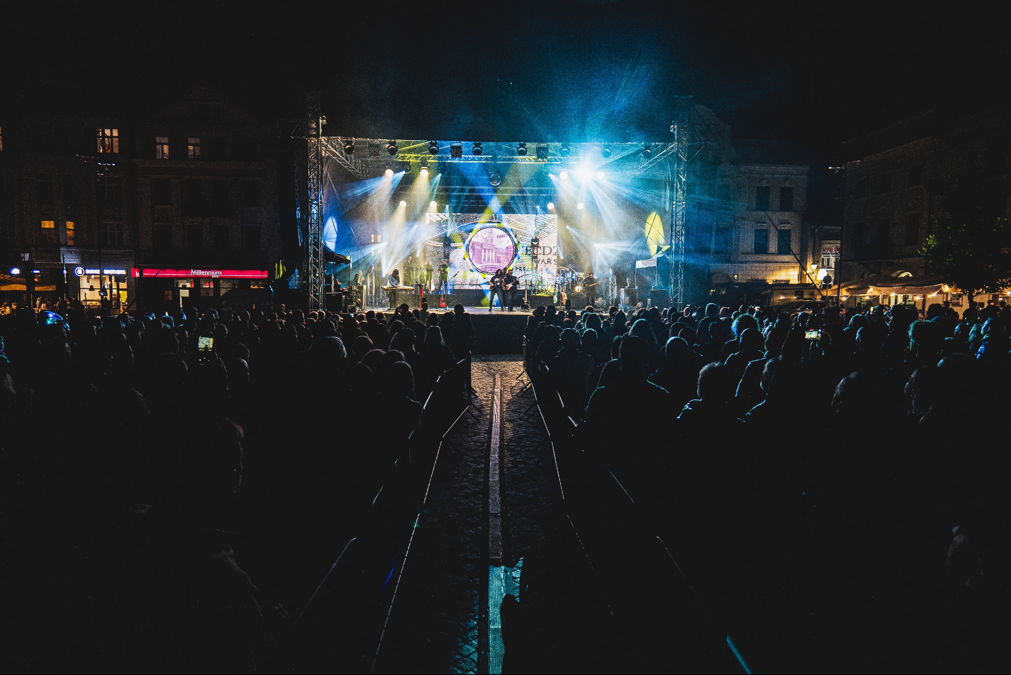 Koncert plenerowy nocą: zespół na scenie oświetlonej reflektorami, tłum widzów w tle, budynki miejskie w oddali.