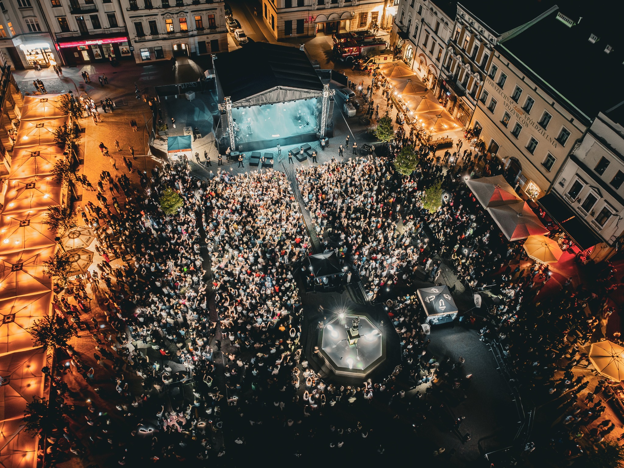 Nocny widok z lotu ptaka na koncert na rynku miejskim, tłum ludzi przed sceną, oświetlona fontanna, budynki w tle.