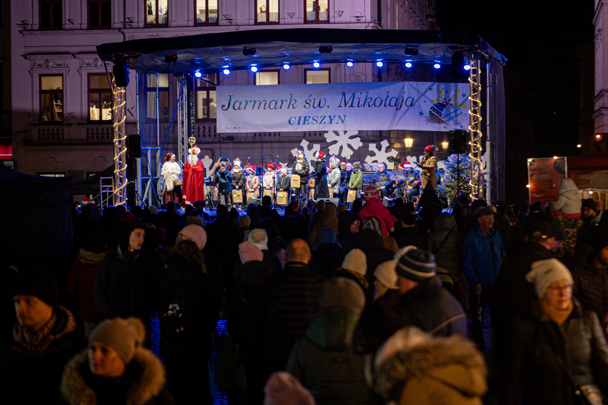 Scena na Jarmarku św. Mikołaja w Cieszynie, na której występują dzieci w czerwonych czapkach i orkiestra dęta, oświetlona niebieskimi reflektorami, przed sceną zgromadzony tłum widzów.